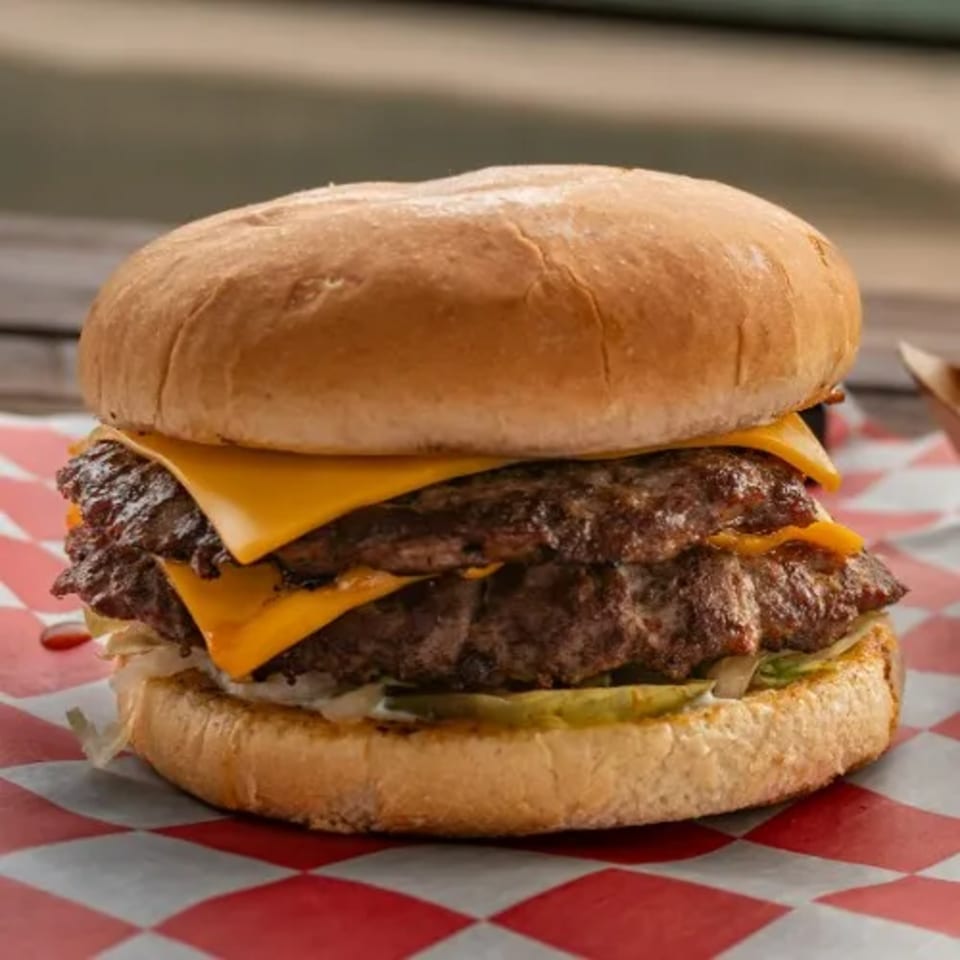 A double cheeseburger from 2-M's Malt N Burger Mart with melted cheese, pickles, and lettuce on a red and white checkered paper.