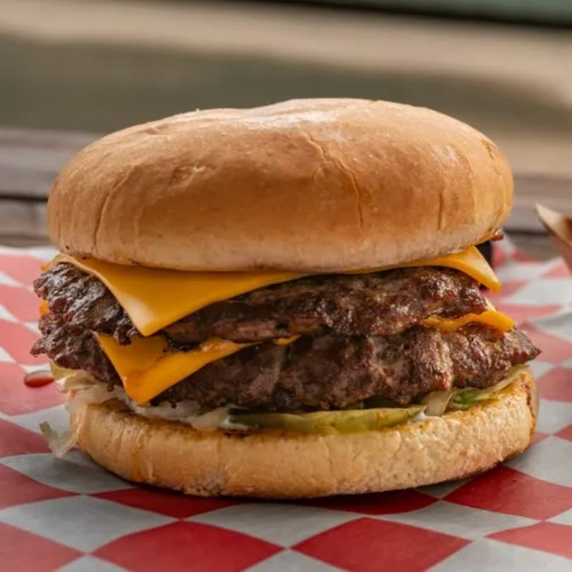 A double cheeseburger from 2-M's Malt N Burger Mart with melted cheese, pickles, and lettuce on a red and white checkered paper.
