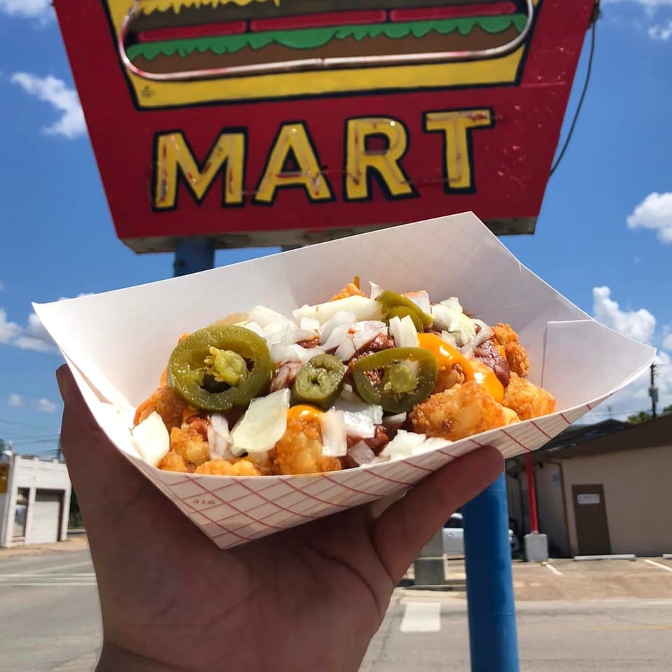 Hand holding loaded tater tots with cheese, chili, onions, and jalapeños, in front of the 2-M's Malt N Burger Mart sign.