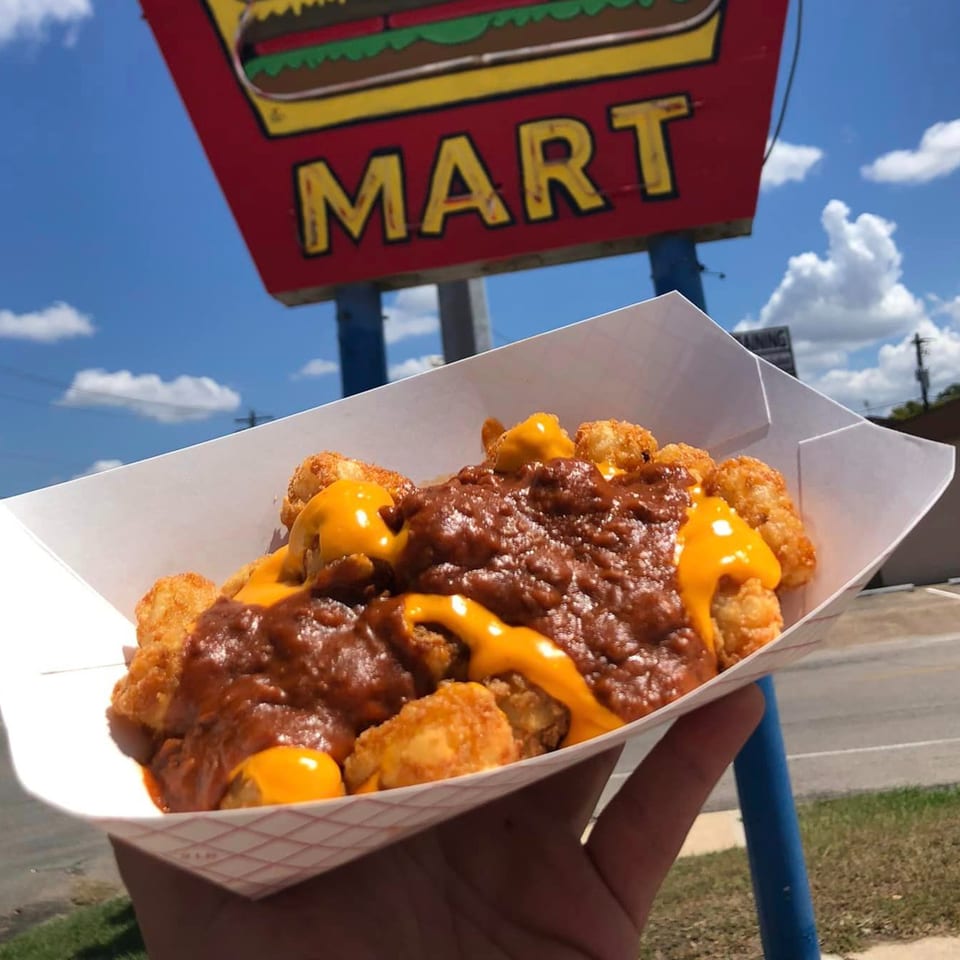 Hand holding a tray of chili cheese tater tots in front of the 2-M's Malt N Burger Mart sign.