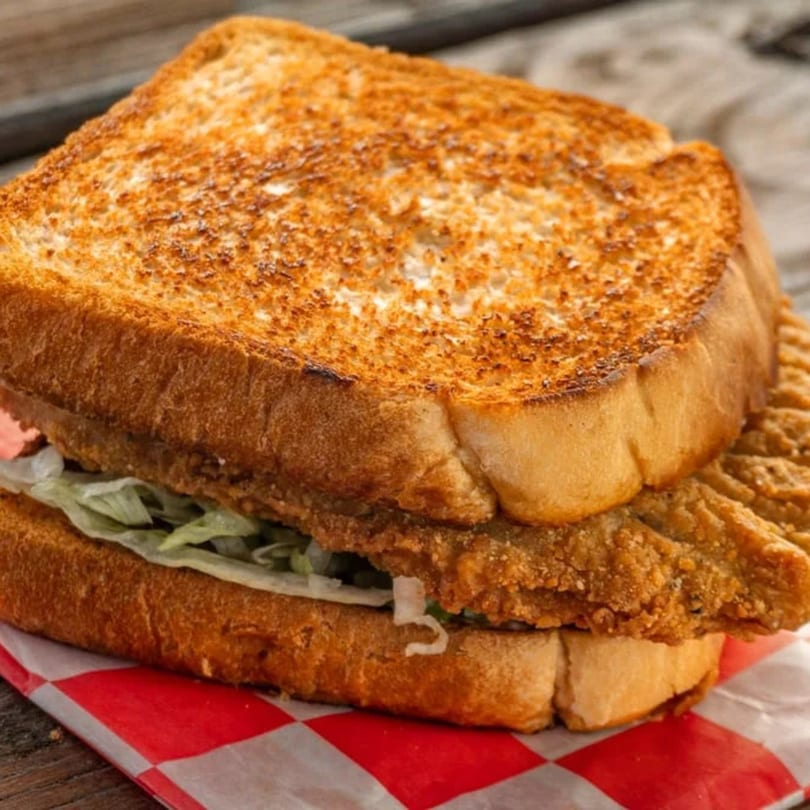 A fried fish sandwich on toasted bread with lettuce, served on red and white checkered paper.