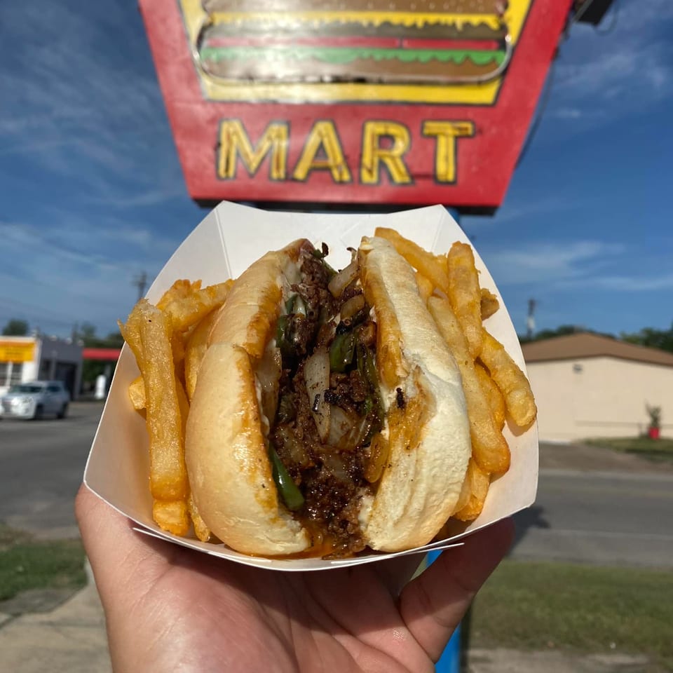 A hand holds a cheesesteak and fries in a paper tray, with the 2-M's Malt N Burger Mart sign in the background.